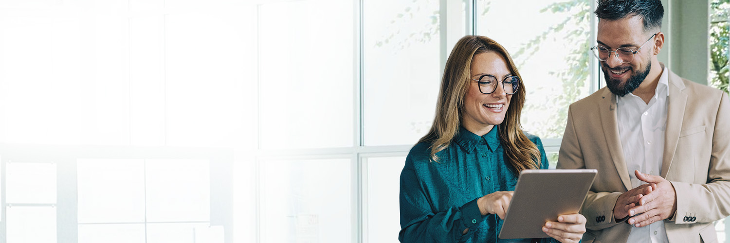 A bearded man wearing professional clothes smiles while standing next to a professional woman wearing glasses as the woman holds a mobile tablet and points to show the man information about HM Insurance Group's Stop Loss and Specialty Insurance solutions.